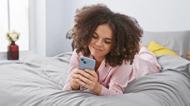 カーリーヘアの陽気なヒスパニック女性はベッドに横たわり スマートフォンは手で 優れたOkサインをジェスチャーし 居心地の良い寝室の背景にフレンドリーな笑顔を点滅させます — ストック動画