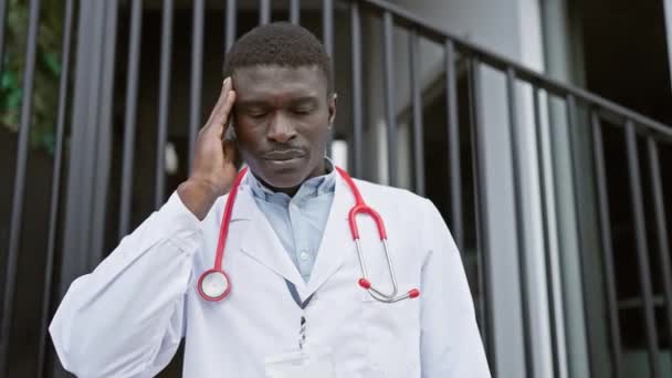 身穿医生制服 手持听诊器站在医院 手扶额头 站在城市环境中的焦虑的非洲男人 — 图库视频影像