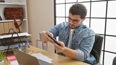 Modern bir ofis ortamında akıllı telefon kullanan gözlüklü ve kulaklıklı genç bir adam.