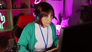 Kulaklıklı yetişkin bir İspanyol kadın geceleri neon ışıklı bir odada bilgisayar kullanıyor.
