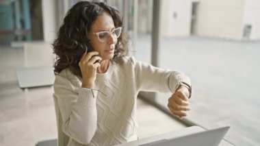 Orta yaşlı bir kadın modern bir ofis ortamında telefonla konuşurken saatini kontrol ediyor..