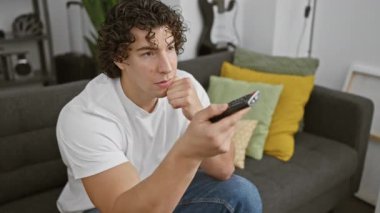 Kıvırcık saçlı genç bir İspanyol evdeki kanepede oturuyor ve elinde düşünceli bir ifadeyle uzaktan kumanda tutuyor..