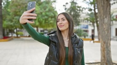 Parka giymiş genç bir kadın akıllı telefonuyla selfie çekiyor, şehir parkında gülümsüyor..