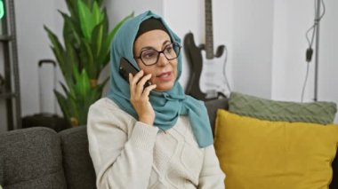 Başörtülü olgun bir kadın kapalı alanda telefonla konuşur, rahat ve modern bir ev ortamı gösterir..