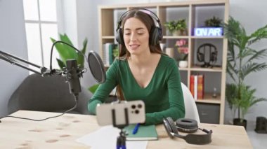 Yeşil bluzlu odaklanmış bir kadın modern bir stüdyoda kulaklık, mikrofon, akıllı telefon ve 'yayında' tabelasıyla bir podcast kaydediyor..