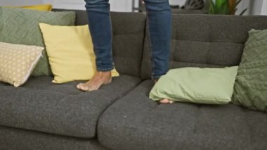 Kot pantolonlu bir adam, modern oturma odasındaki renkli yastıklarla gri bir koltukta yalınayak yürür..