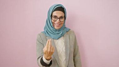 Mutlu, orta yaşlı İspanyol kadın tesettürlü, mutlu, pembe izole bir arka plana karşı el hareketleriyle buraya davet ediyor..