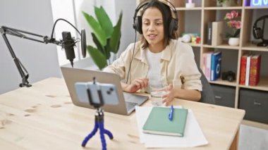 Podcast stüdyosunda akıllı telefon ve mikrofonla su içen İspanyol kadın..