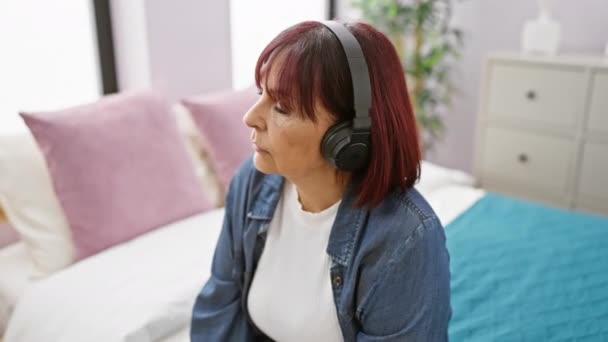 成熟したヒスパニック女性は自宅で彼女の居心地の良い寝室でリラックスしてヘッドフォンで音楽に耳を傾ける — ストック動画