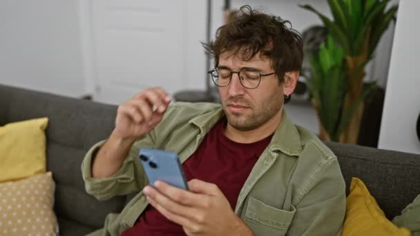 一个留着胡子的紧张的年轻人在屋里沙发上用智能手机 — 图库视频影像