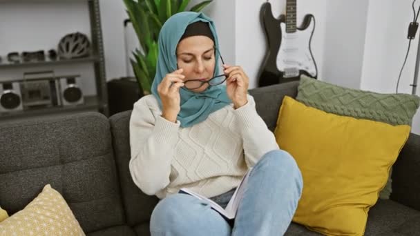 Dojrzała Kobieta Okularach Hidżab Czyta Książkę Przytulnym Salonie Gitarą Tle — Wideo stockowe