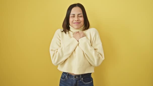 陽気な若いヒスパニック系の女性がセーターを着用し 胸に感謝の意を表し 美しく微笑んだ 活気に満ちた黄色の隔離された背景に誇らしげに立って 健康を放射する — ストック動画