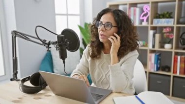 Latin kökenli olgun kadın podcaster ev telefonunda dizüstü bilgisayar ve mikrofonla konuşuyor..