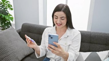Genç bir İspanyol kadın, modern bir oturma odasında hamilelik testi yaparken telefonuna gülümsüyor..