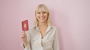 Gülümseyen, beyaz, sarışın bir kadın, Danimarka pasaportunu pembe arka planda tutuyor. Seyahat ve kimliği simgeliyor..
