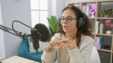 Modern radyo stüdyosunda kulaklıklı, gülümseyen orta yaşlı İspanyol kadın mikrofona konuşuyor.