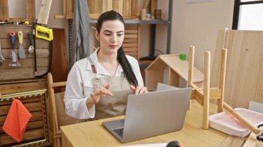 Genç bir İspanyol kadın marangozluk atölyesinde dizüstü bilgisayarla meşgul oluyor, modern, çekici bir girişimciyi örnek olarak gösteriyor. kapalı yaratıcı stüdyo alanında..