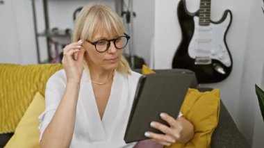 İçerideki sarı koltukta tablet kullanan odaklanmış gözlüklü bir kadın. Arka planda gitar var.