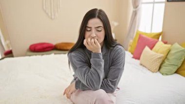 Endişeli, genç ve güzel bir İspanyol kadın gergin bir şekilde tırnak yiyor, pijama giyiyor, stresli görünüyor yatak odasında oturuyor - bir ev stresi sorunu portresi.