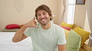 Neşeli, genç, beyaz bir adam mutlu bir şekilde, rahat yatak odasında parmaklarıyla telefon işareti yapıyor, pozitifliğini ve dostça iletişim kavramını dile getiriyor..