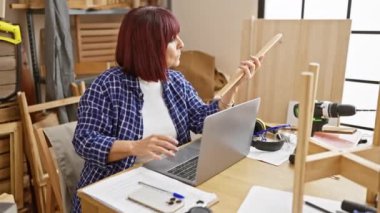 Olgun kadın, marangozluk atölyesinde dizüstü bilgisayar ve aletlerle ahşabı inceliyor.