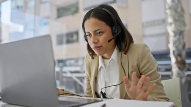 Kulaklıklı genç İspanyol kadın şehir manzaralı modern ofiste dizüstü bilgisayarla çalışıyor..
