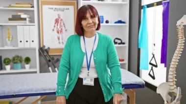 Orta yaşlı İspanyol kadın profesyonel olarak bir rehabilitasyon kliniğinde pano, anatomi posteri ve fizyoterapi ekipmanıyla duruyor.
