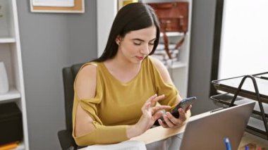 Ofiste çalışan genç bir Latin kadın masasında otururken telefonunu kullanıyor..