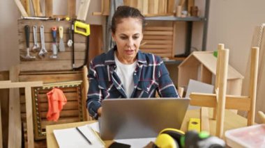 Orta yaşlı, kıvırcık saçlı bir İspanyol kadın marangozluk atölyesinde rahatlıyor, dizüstü bilgisayarla çalıştıktan sonra esniyor.