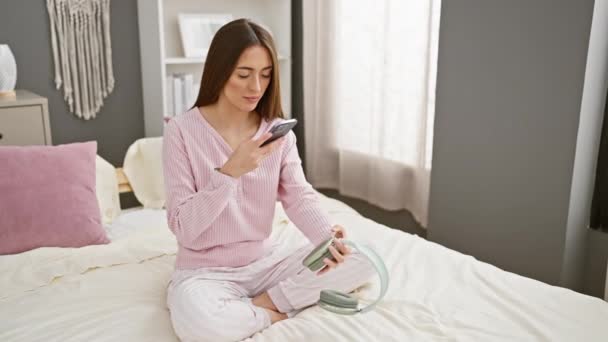 睡衣女人穿着睡衣在卧室的床上用智能手机操作耳机的女人 — 图库视频影像