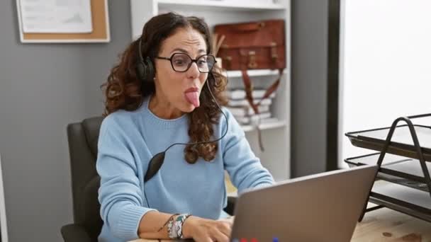 彼女はヘッドフォンでラップトップで動作するように カーリーヘアの成熟した女性は 現代のオフィスの設定で彼女の舌を遊び出します — ストック動画
