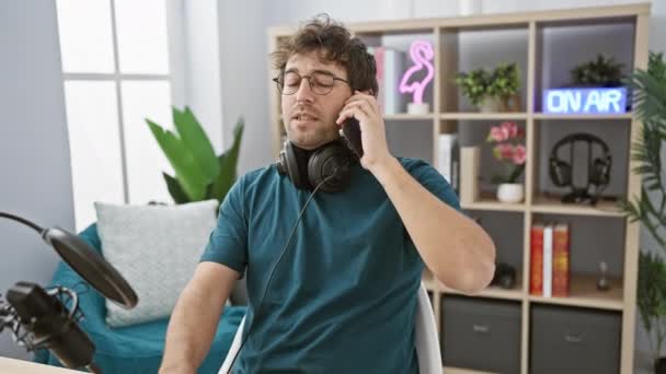 一位留着胡子的年轻人在电话里交谈 并在一个带有 标志的广播演播室里喝咖啡 — 图库视频影像