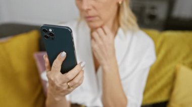 Beyaz gömlekli tefekkür eden sarışın bir kadın arka planda sarı kanepesi olan kapalı bir yerde akıllı telefon kullanıyor..