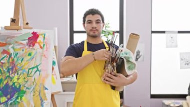 Sarı önlüklü neşeli bir adam parlak bir stüdyoda sanat malzemeleri tutuyor..