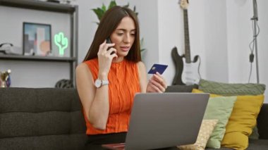 Bir kadın elinde kredi kartıyla akıllı telefonuyla konuşur. Modern bir oturma odasında dizüstü bilgisayara bakar..