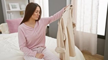 Pijamalı bir kadın, temiz bir yatak odasında yatağında bej renkli bir trençkotu inceliyor.