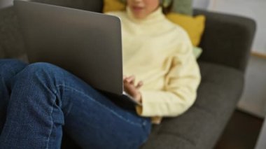 Sarı kazaklı İspanyol kadın içeride gri koltukta otururken dizüstü bilgisayar kullanıyor, kulaklık takıyor..