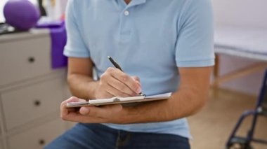 Rehabilitasyon kliniğinde panoya yazı yazan yakışıklı sakallı bir adam. Mavi bir polo giyiyor ve gülümsüyor..