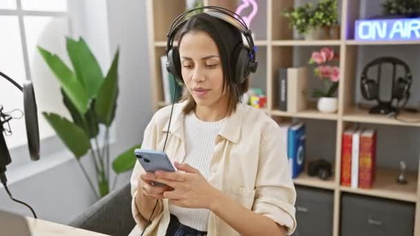 一位戴着耳机的年轻女子在一个配备了麦克风和装饰的现代广播演播室里使用智能手机 — 图库视频影像