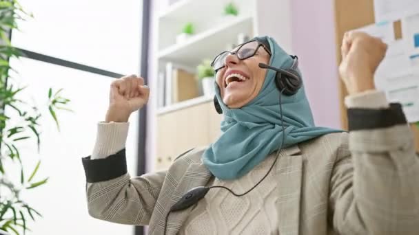 Dojrzała Kobieta Hidżabie Radośnie Świętuje Podniesionymi Pięściami Biurze Słuchawkach — Wideo stockowe