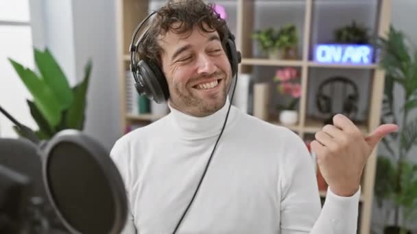 ラジオスタジオで陽気な若いヒスパニック男性 親指で側面を指し ヘッドフォンでロックアウト — ストック動画