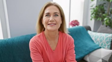 Evdeki kanepede oturan neşeli, orta yaşlı sarışın kadın, kendine güvenle dişlek bir gülümseme sergiliyor. Sevinçli ve neşeli, o mutlu tavırlarıyla pozitifliğini sergiliyor..