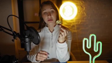 Genç bir kadın, modern finans eğilimlerini sembolize eden neon ışıklı podcast stüdyosunda bir bitcoin 'i inceliyor..