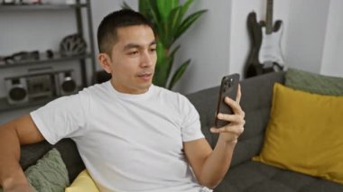 Güzel dekore edilmiş bir oturma odasındaki kanepede uzanırken akıllı telefon kullanan sıradan bir genç..