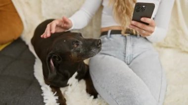 Sarışın kadın rahat oturma odasında akıllı telefon kullanırken köpeğini okşuyor.