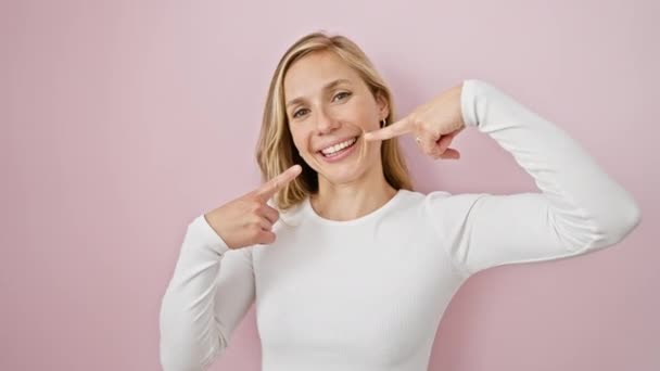 魅力的な若いブロンドの女性は ピンクの背景の上に立って 歯の健康をオフに示す彼女の完璧な歯を元気づけます — ストック動画