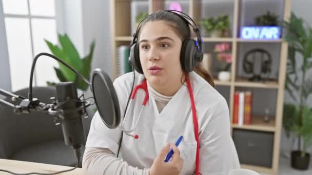 ヘッドフォンを持つ若いヒスパニック系女性がラジオスタジオでマイクに話しかけ プロのオーディオ機器とエアサインを披露する — ストック動画