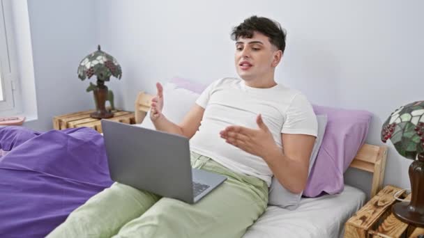若い男は カラフルな寝室に座っている間 ラップトップ上のビデオ通話を通じて誰かと交流します — ストック動画