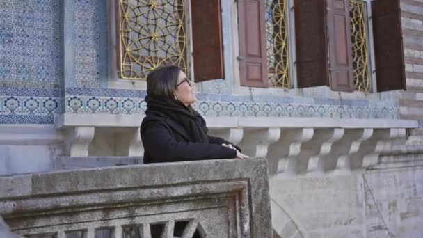 笑顔の女性が眼鏡をかけ 黒いスカーフがイスタンブールのトップカピ宮殿に立っており 歴史的なオスマン建築を展示しています — ストック動画