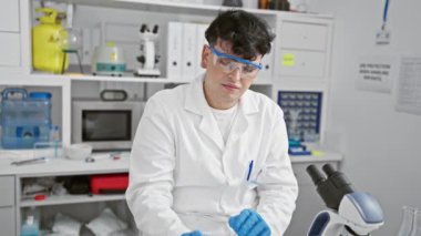 Laboratuvar önlüklü bir adam, bilimsel araştırmaları gösteren mikroskoplar ve deney tüpleriyle modern bir laboratuvarda test tüpünü inceliyor..